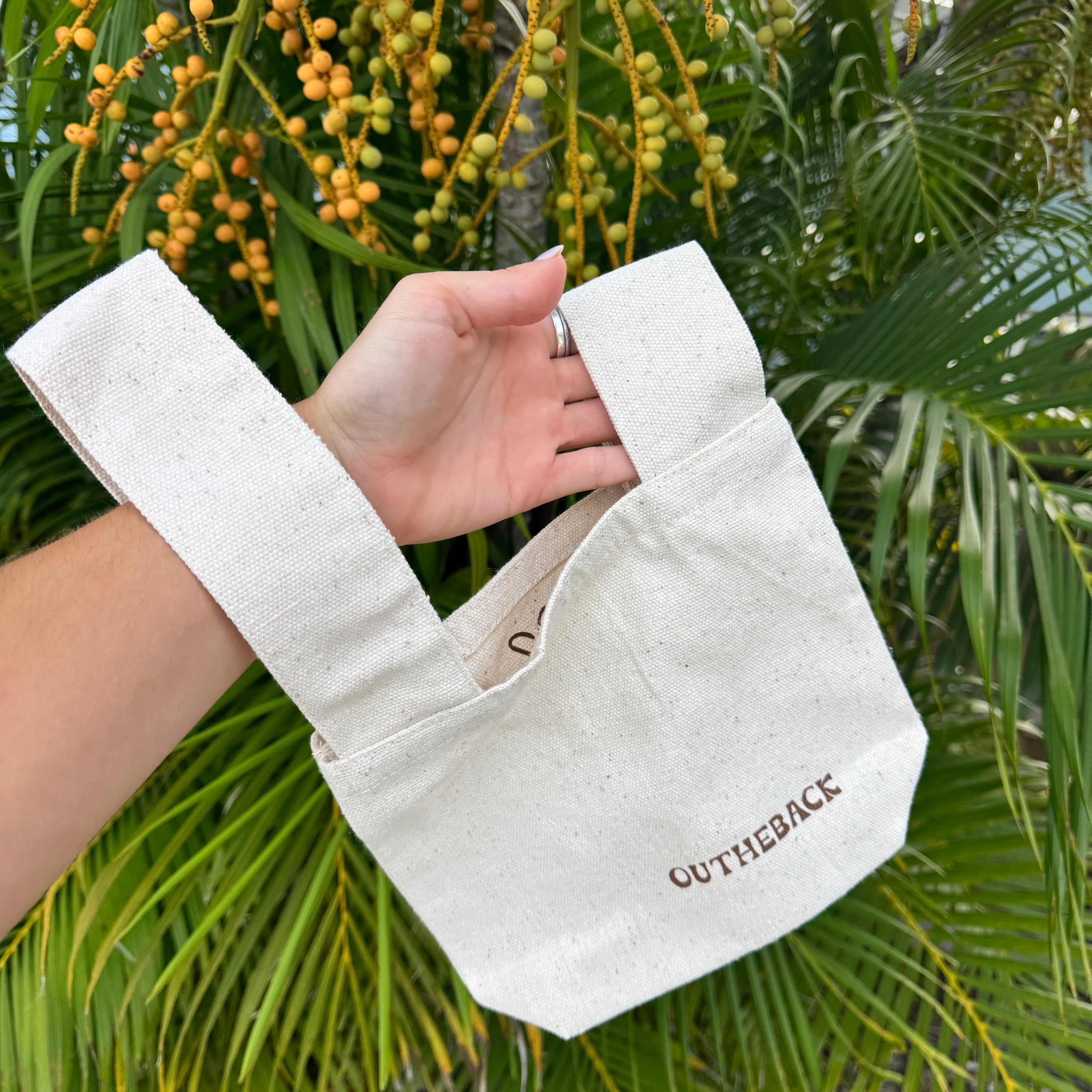 A hand holding a cream-colored canvas tote bag with the brand name 'OUTHEBACK' printed in brown on the front, set against a background of green palm leaves and yellow-orange hanging berries.