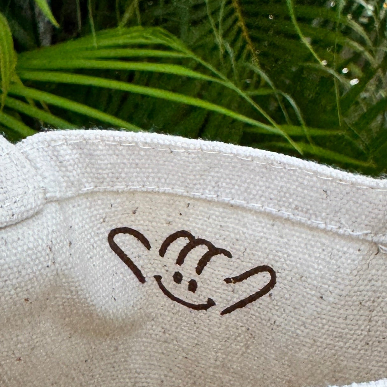 A close-up of a natural beige canvas tote bag held by a hand, featuring a brown hand gesture logo printed inside. The background shows lush green tropical foliage.