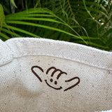 A close-up of a natural beige canvas tote bag held by a hand, featuring a brown hand gesture logo printed inside. The background shows lush green tropical foliage.