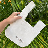 A hand holding a light beige canvas tote bag with the brand name 'OUTHEBACK' printed in dark brown on the front, set against a background of green palm leaves and yellow-orange palm fruits.