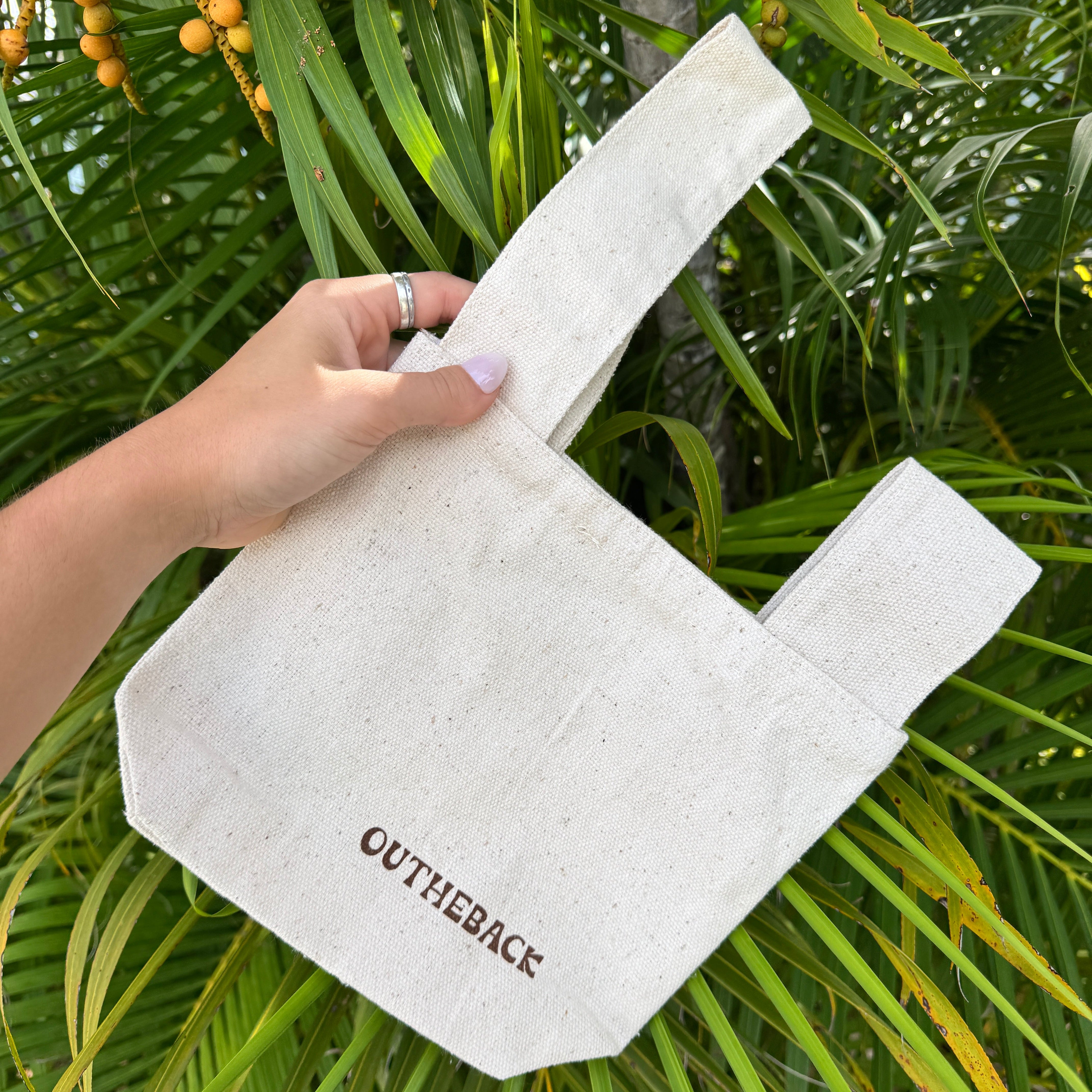 A hand holding a light beige canvas tote bag with the brand name 'OUTHEBACK' printed in dark brown on the front, set against a background of green palm leaves and yellow-orange palm fruits.