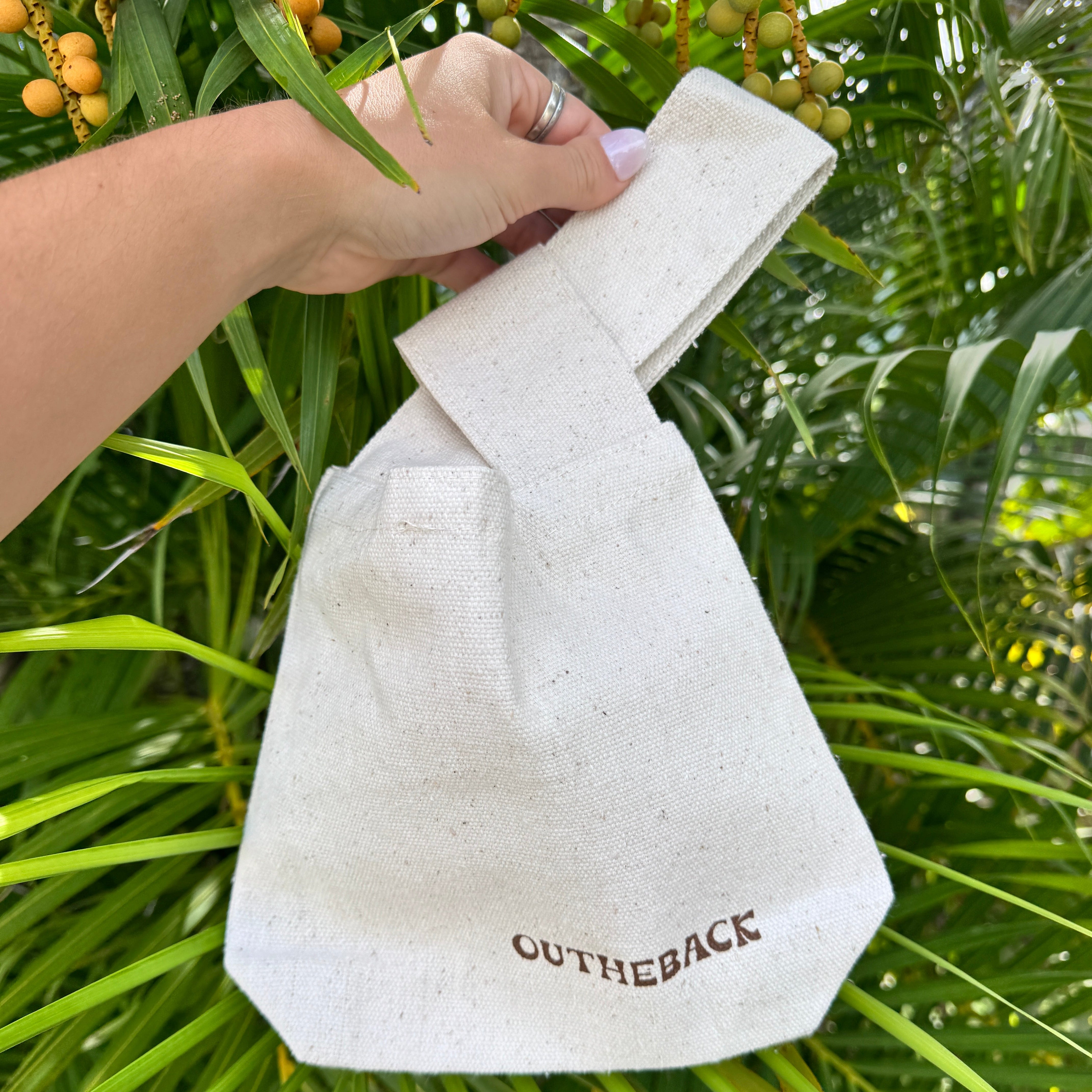 A hand holding a light beige canvas tote bag with a twisted top handle, featuring the brand name 'OUTHEBACK' printed in brown on the front, set against a background of green palm leaves and small yellow-orange berries.