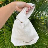 A hand holding a small off-white canvas tote bag with a loop handle, featuring the brand name 'OUTTHEBACK' printed in brown near the bottom, set against a background of green palm leaves.