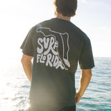Person wearing a black t-shirt with 'Surf Florida' design by the ocean