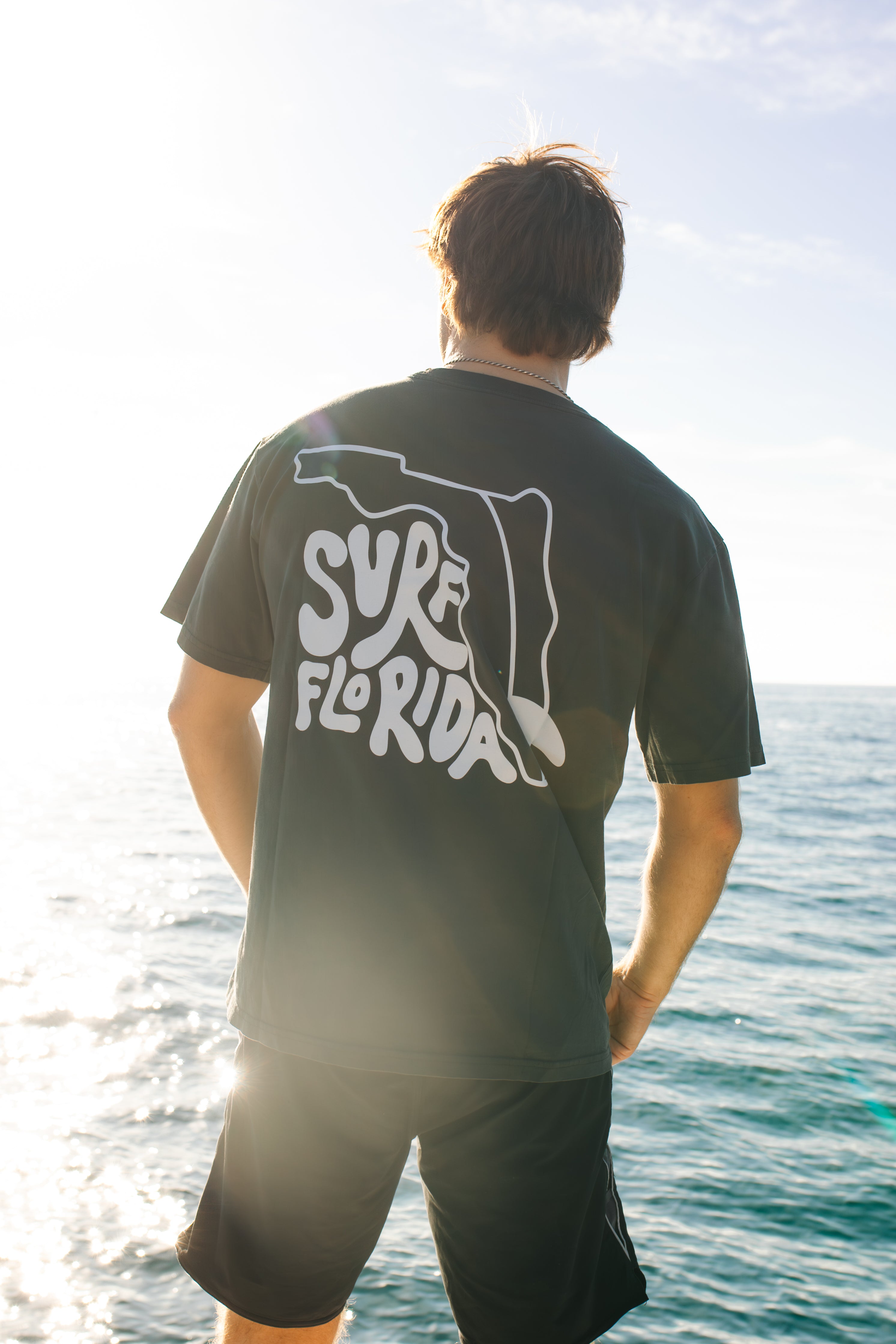 Person wearing a black t-shirt with 'Surf Florida' design by the ocean