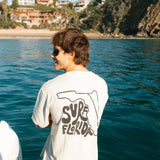 Person wearing a 'Supreme Florida' shirt by a body of water with houses on a hillside.