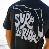 Person wearing a black t-shirt with 'Surf Florida' design, standing by water.