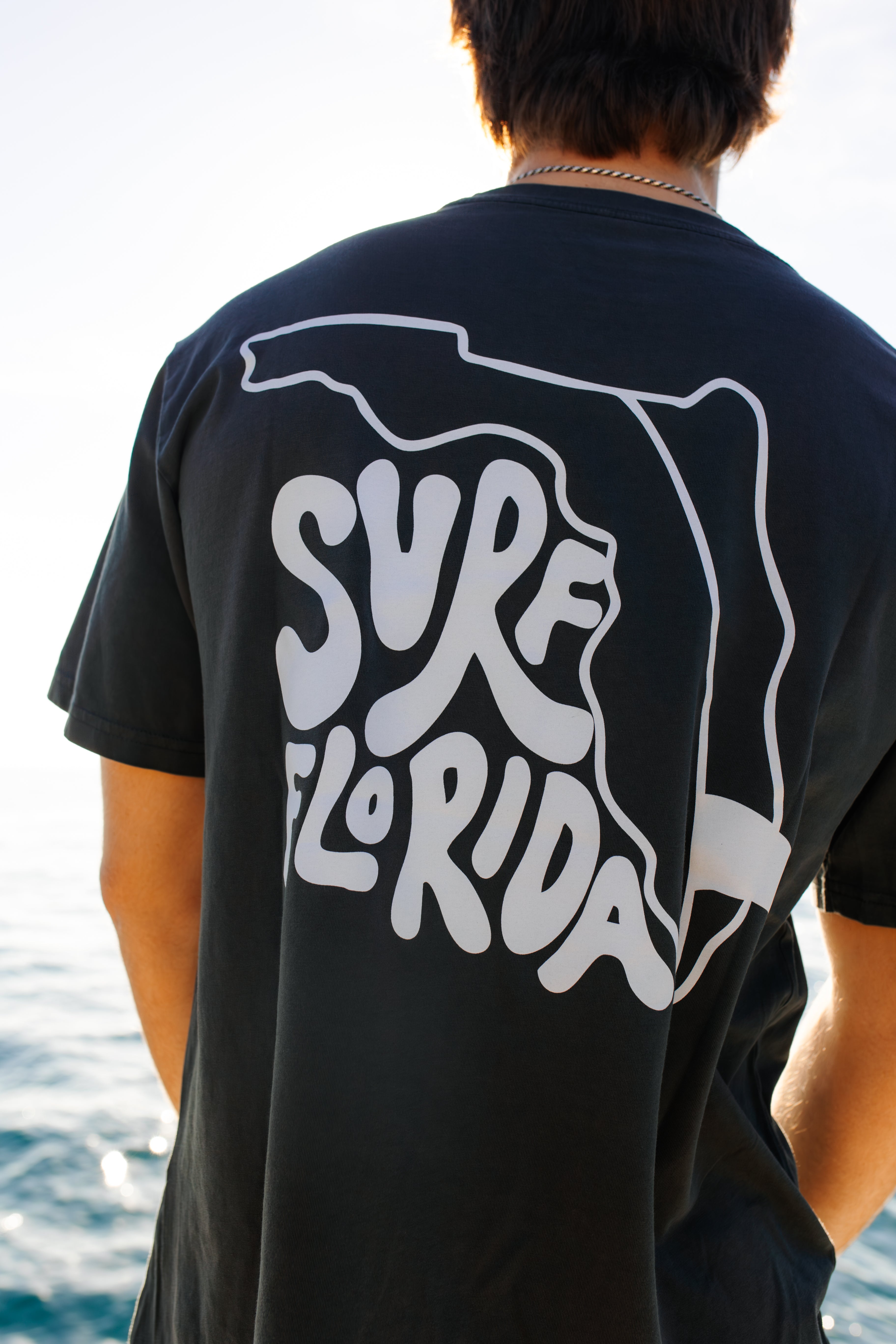 Person wearing a black t-shirt with 'Surf Florida' design, standing by water.