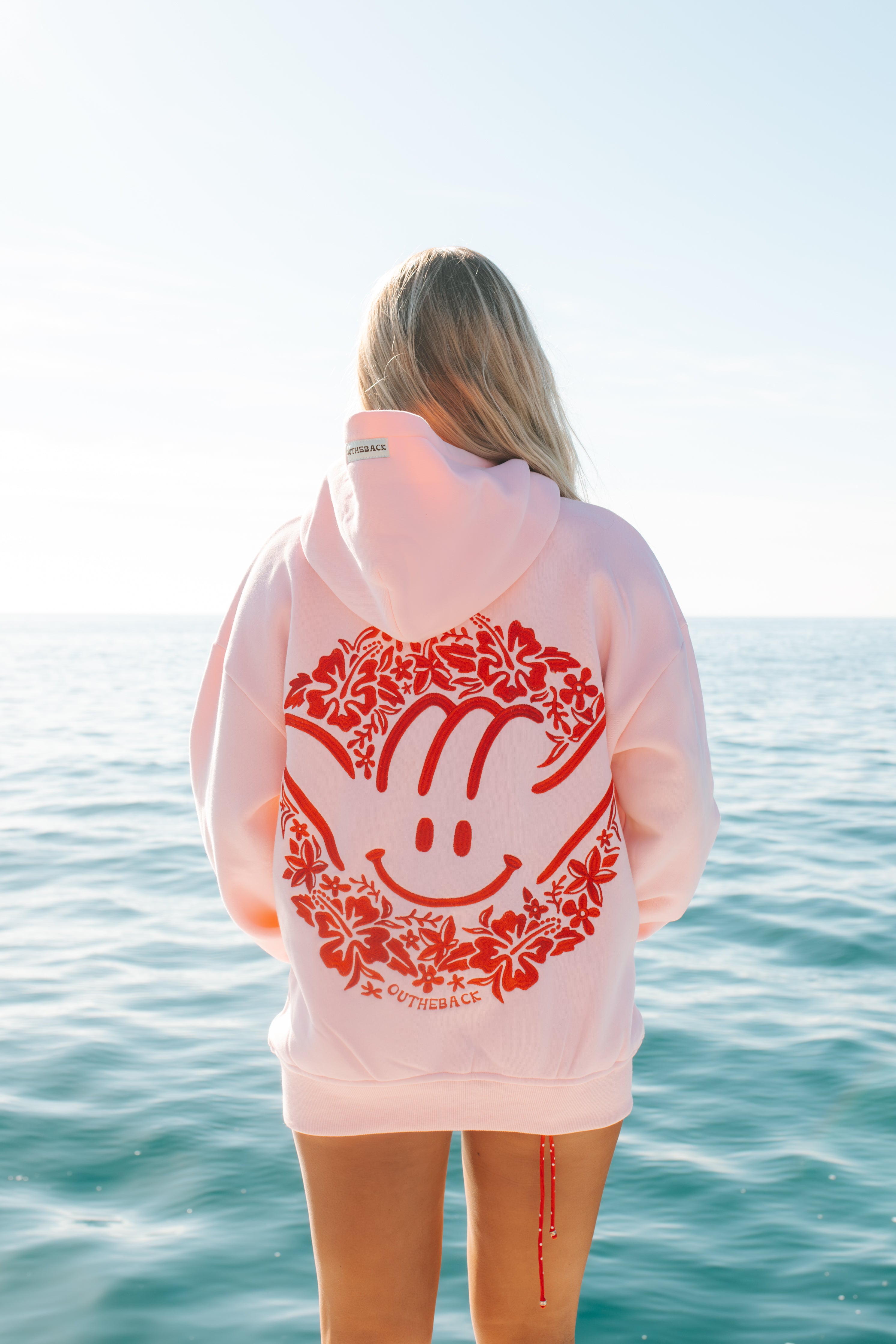 Person wearing a pink hoodie with a red design by the ocean