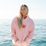 Person wearing a pink hoodie with a logo, standing in front of a body of water.
