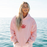 Person wearing a pink hoodie with a logo, standing by the ocean.