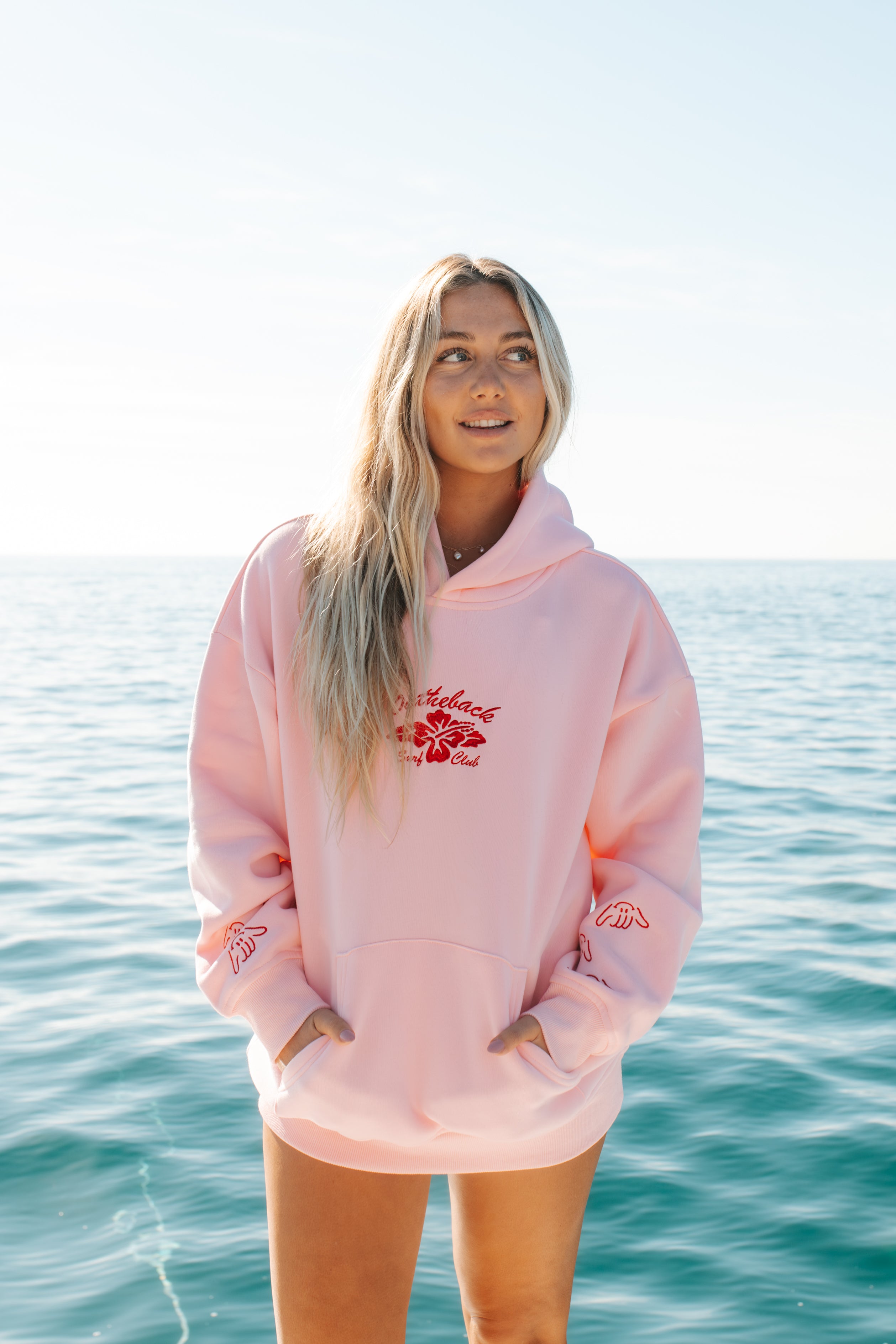 Person wearing a pink hoodie with a logo, standing by the ocean.