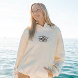 Woman wearing a cream-colored hoodie with a logo, standing by the ocean.
