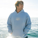 Man wearing a light blue hoodie with a logo, standing by the water.