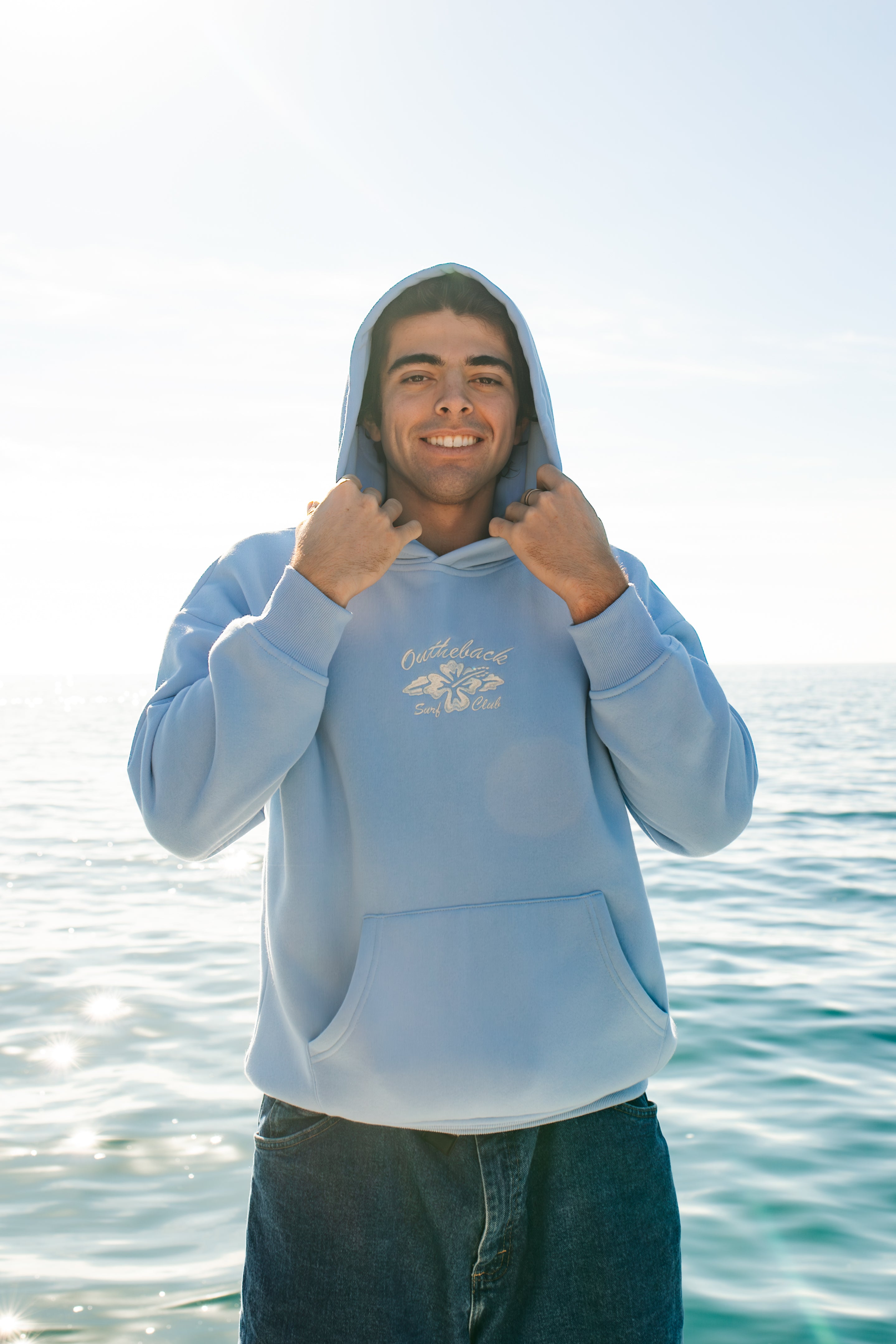 Person wearing a light blue hoodie with a logo, standing by the water.