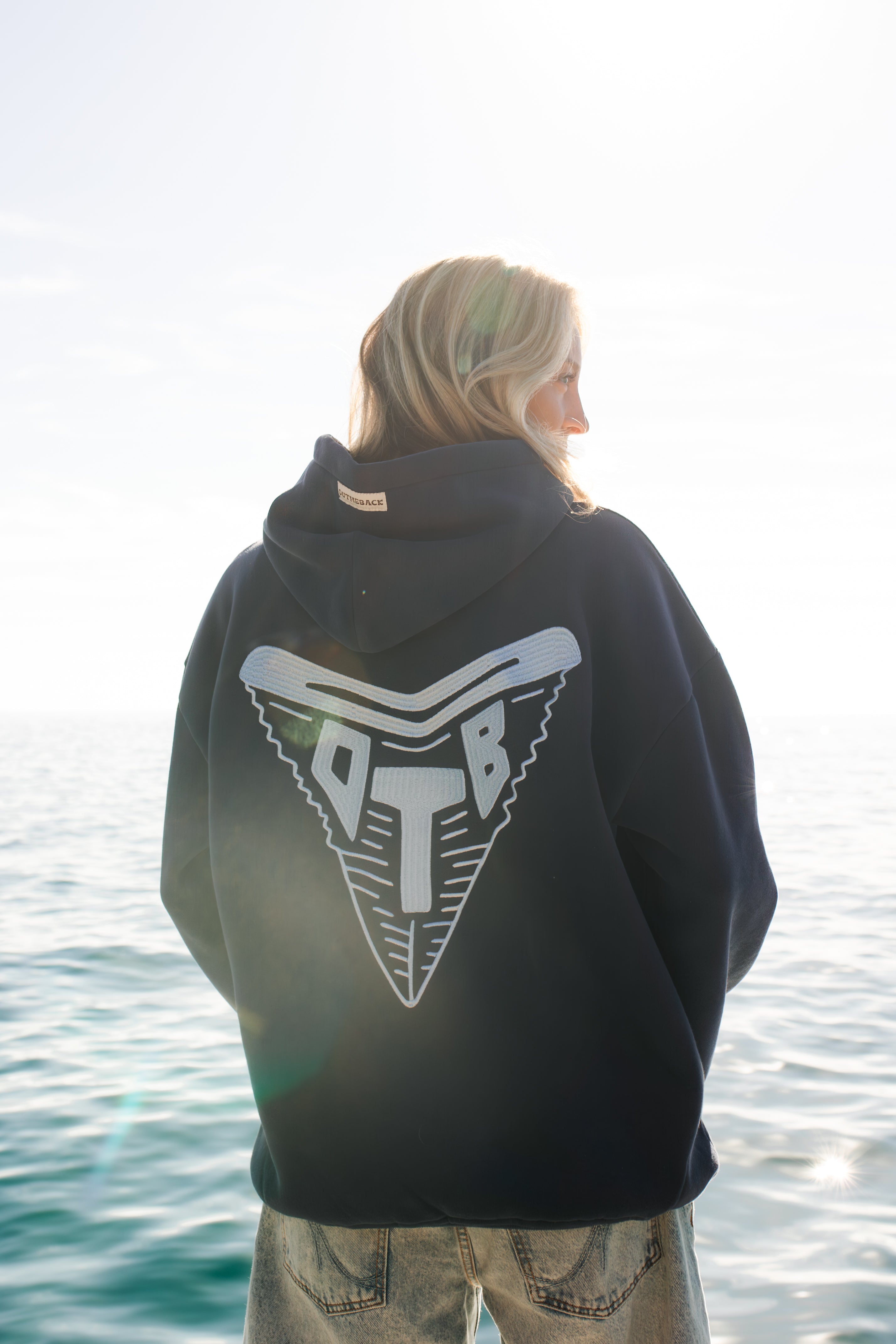 Person wearing a navy hoodie with a shark tooth logo by the water