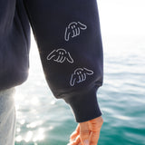 Person wearing a navy blue sweatshirt with Shaka hand designs by a body of water.