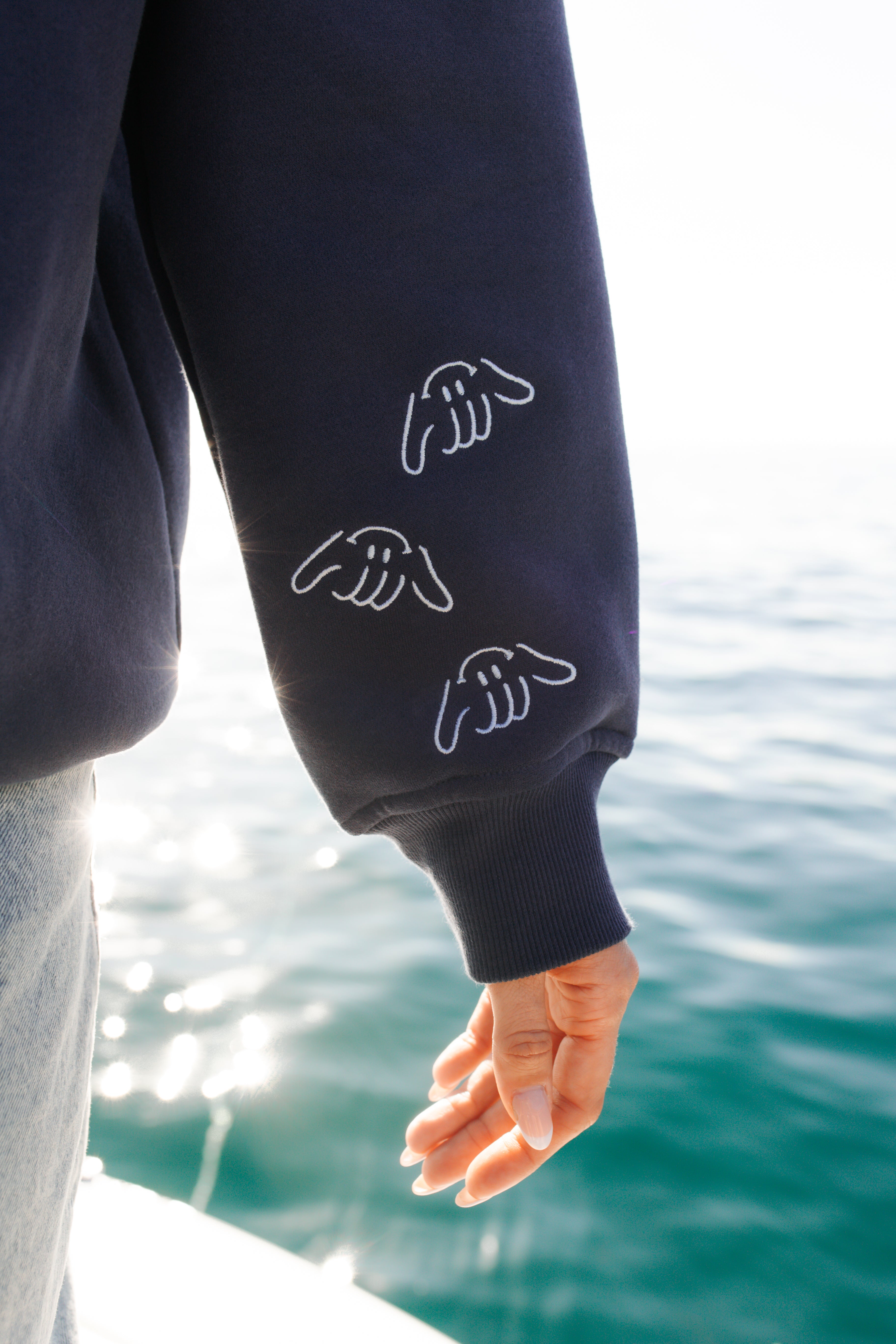 Person wearing a navy blue sweatshirt with Shaka hand designs by a body of water.