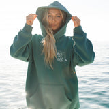 Person wearing a green hoodie with a logo, standing by the water.