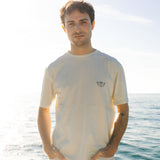 Man wearing a light-colored t-shirt with a logo standing by the ocean.