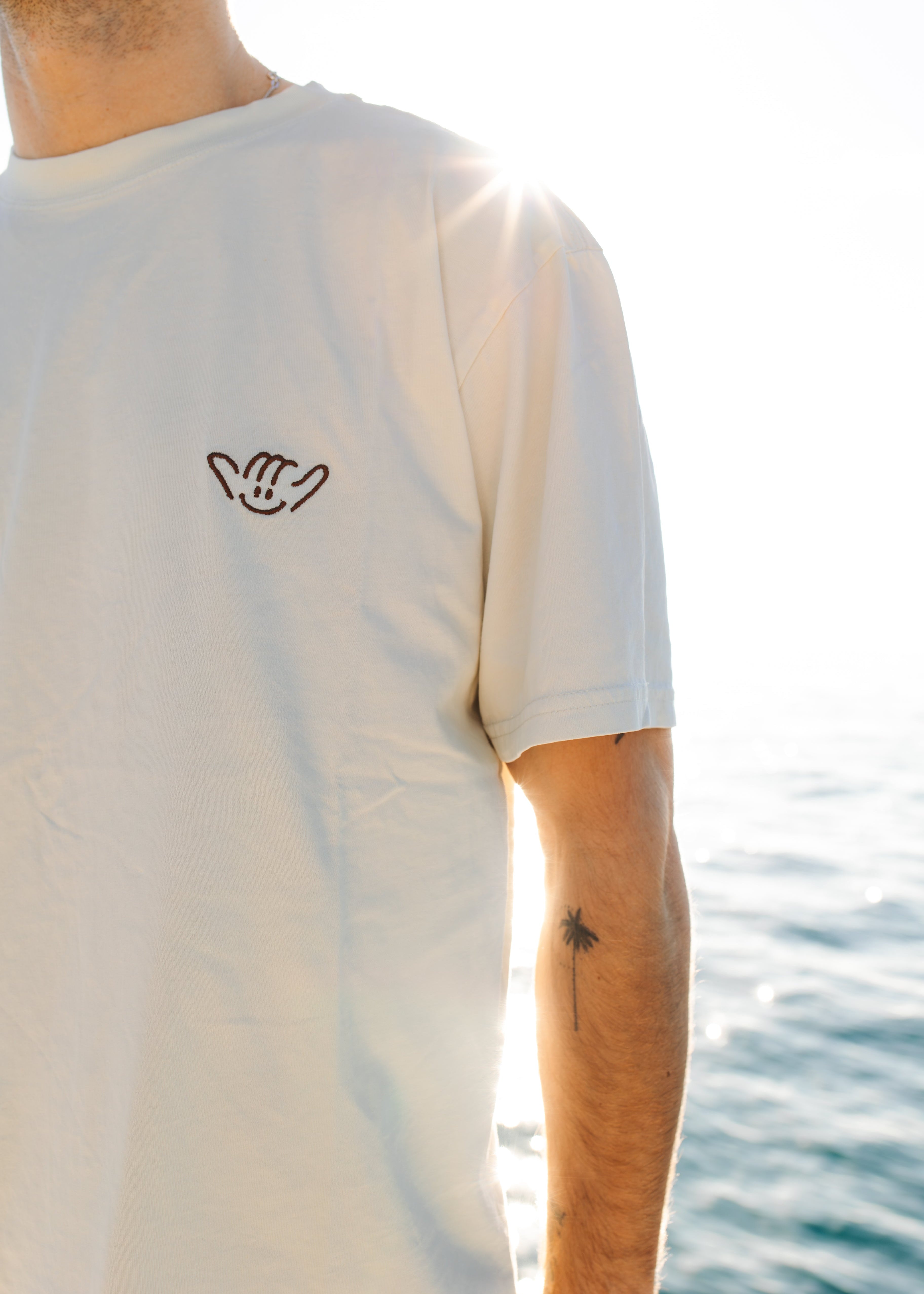 Person wearing a white t-shirt with a small logo, standing by water.