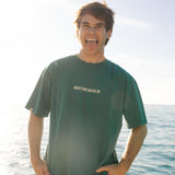 Person wearing a green t-shirt with 'Duthie Back' logo standing by the ocean.