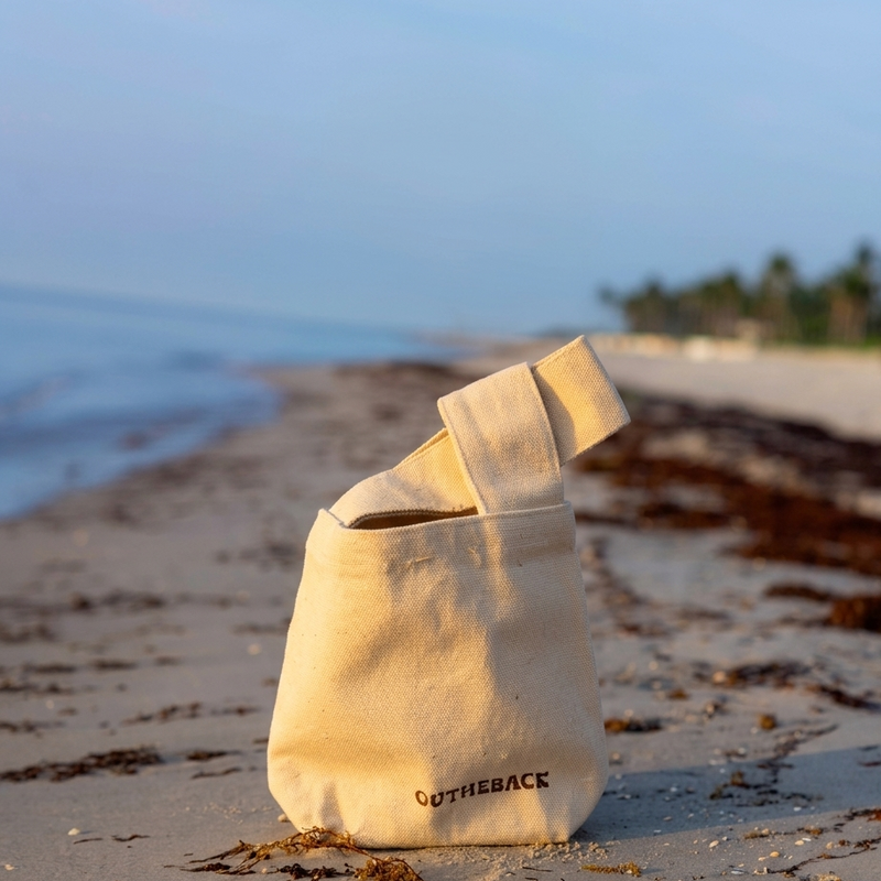 A beige canvas tote bag with black 'OUTHEBACK' branding sits on a sandy beach with the ocean and shoreline blurred in the background.